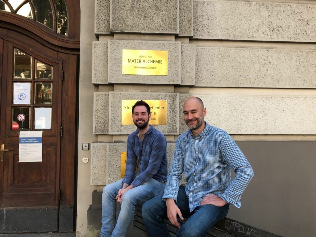 Mitchell Jones und Alexander Bismarck sitzen vor dem Institut für Materialchemie. Beide tragen Jeans und blaue Hemden und lächeln in die Kamera.