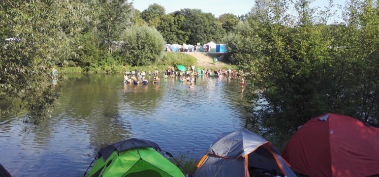 Am Ufer des Flusses stehen mehrere Zelte. Im Fluss sieht man ungefähr vierzig Menschen.