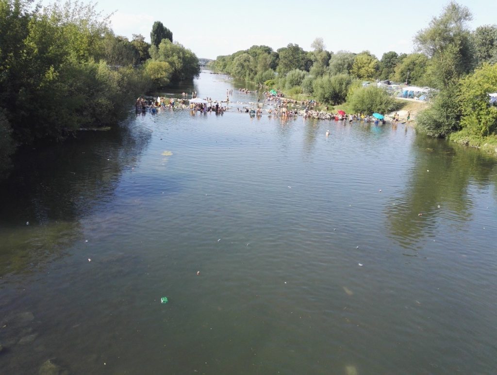 Ein gerader Blick den Fluss entlang, im Hintergrund vergnügen sich Menschen im Wasser. An den Ufern des Flusses stehen Bäume und Sträucher.