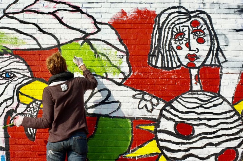 Eine Person mit kurzen Haaren und weißen Handschuhen malt ein Bild an eine Backsteinwand.