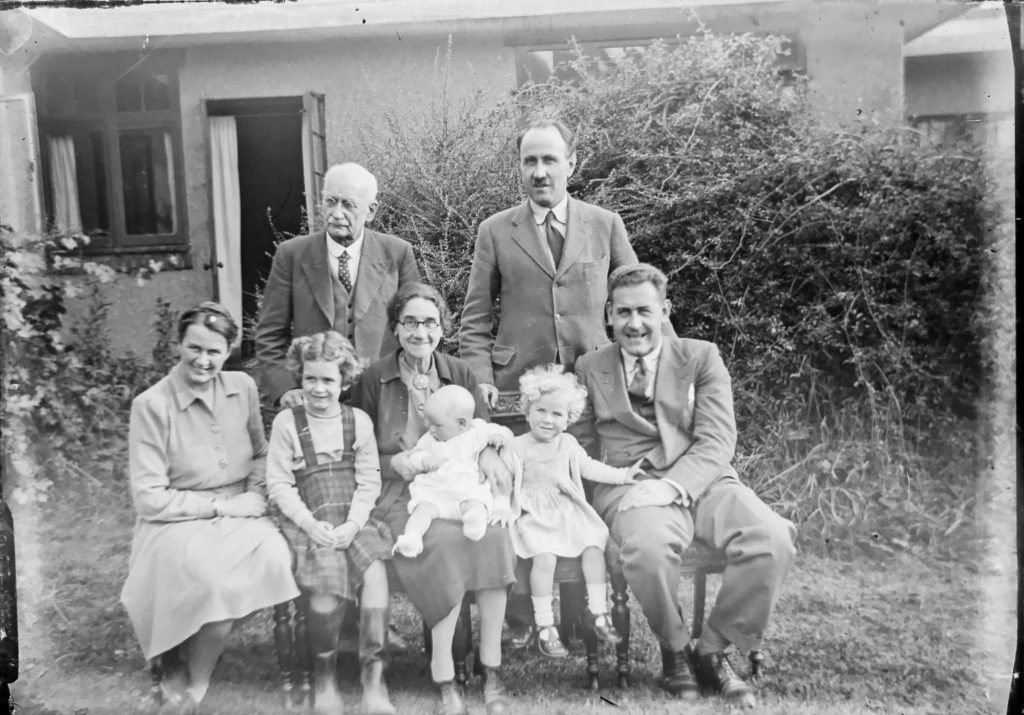 Ein Familienporträt in Schwarz-Weiß. Drei Erwachsene und drei Kinder sitzen auf einer Bank vor einem Haus, dahinter stehen zwei Erwachsene.