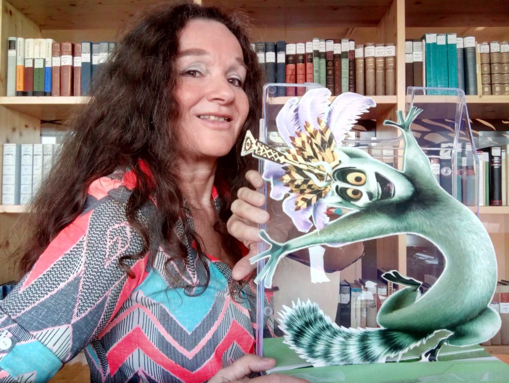 Gerda Moser steht für einem Bücherregal und hält ihr Büromaskottchen, einen Ausdruck von King Julien aus dem Film Madagascar, in die Höhe.