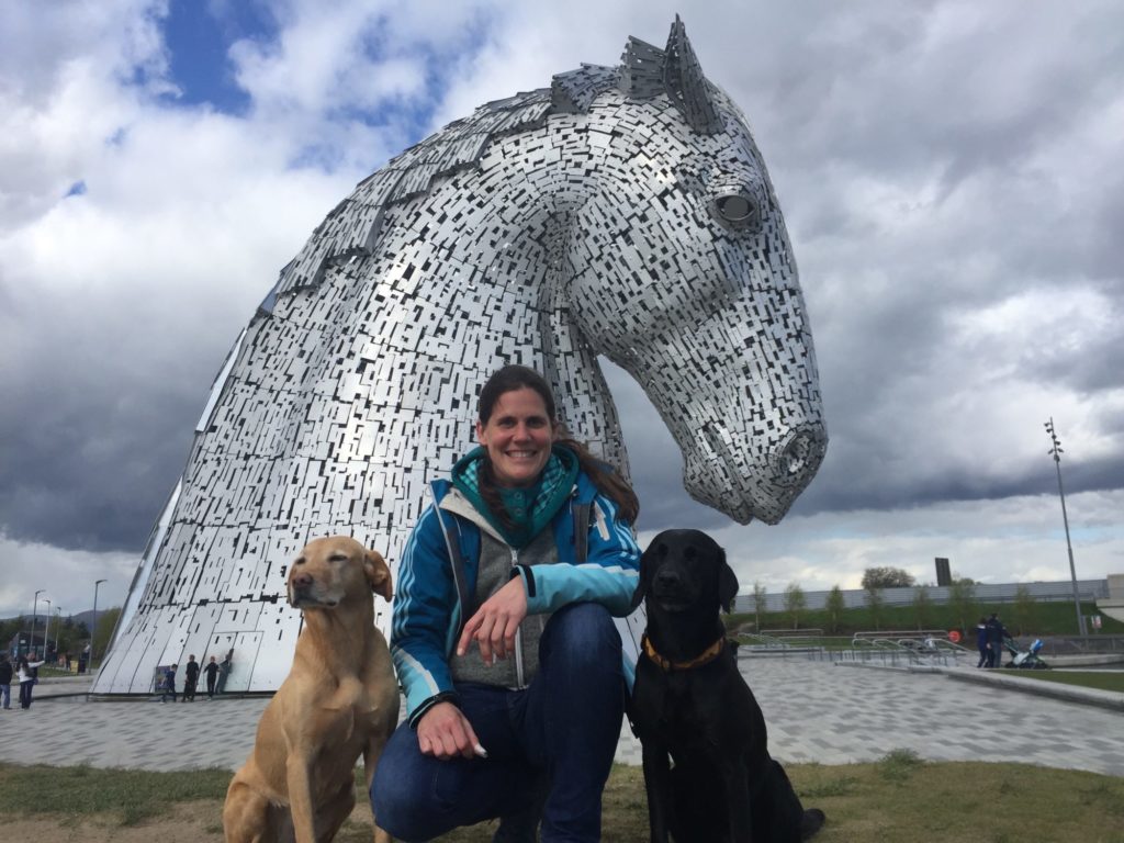 Nadja Affenzeller posiert vor einer großen, silbernen Pferdekopfstatue. Rechts und links von ihr sitzt jeweils ein Hund.