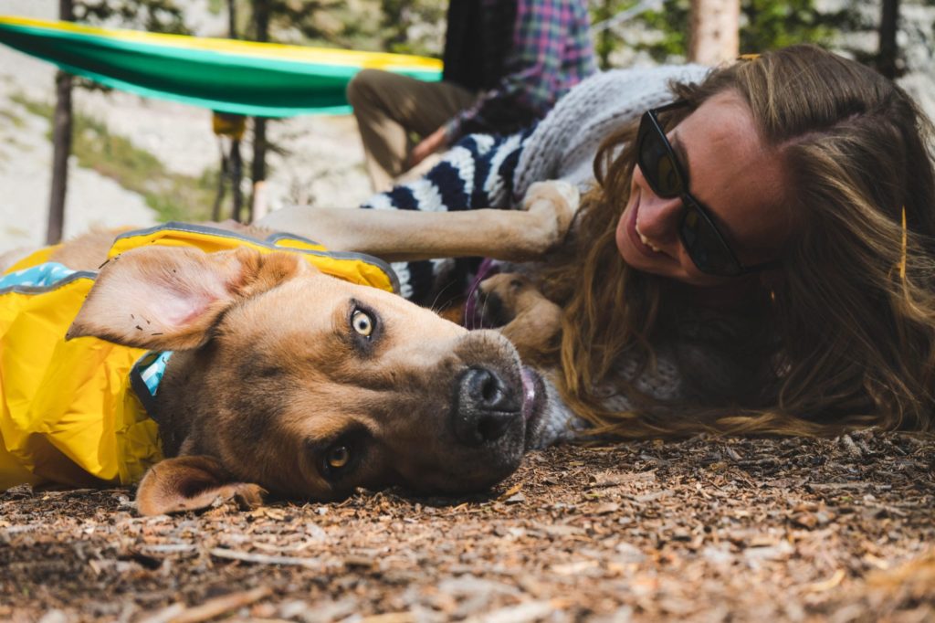 Ein Hund und eine Person mit langen Haaren und einer Sonnenbrille liegen nebeneinander am Boden.