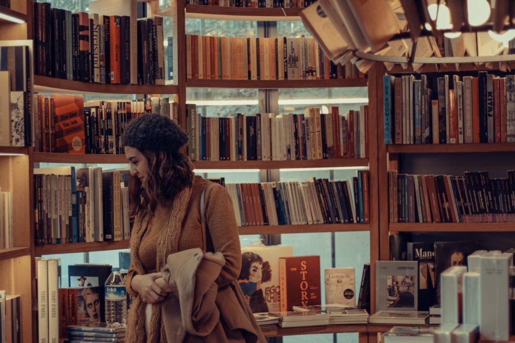 Ein Person sieht sich Bücher in einem Buchladen an. Sie trägt einen braunen Mantel, eine Strickmütze und hat lange Haare.