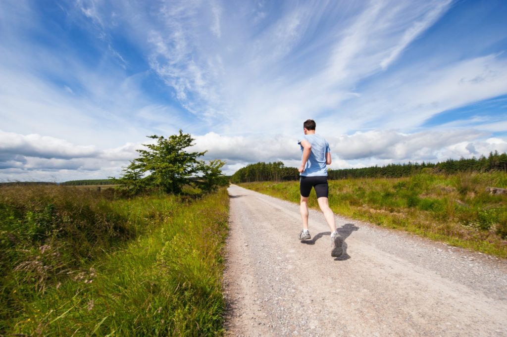 Eine Person in schwarzen Shorts und einem hellblauen T-Shirt läuft einen Weg zwischen zwei Wiesen entlang.