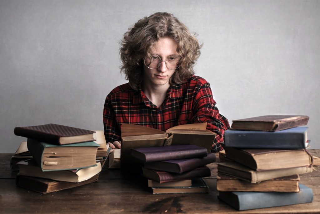 Eine Person mit Locken und einer Brille sitzt an einem Tisch vor Stapeln alt aussehender Bücher.