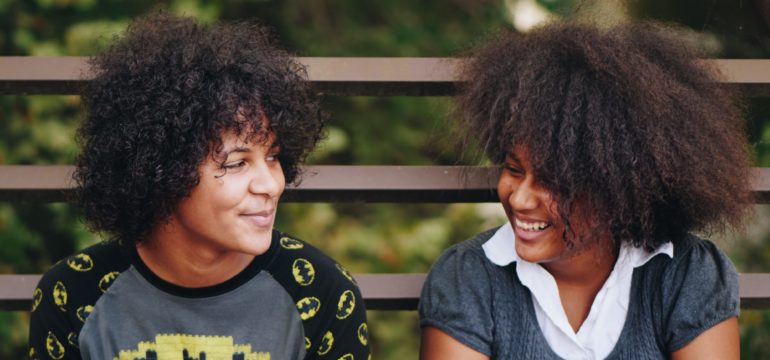 Zwei Personen mit dunklen, krausen Haaren sitzen auf einer Bank und sehen einander an.
