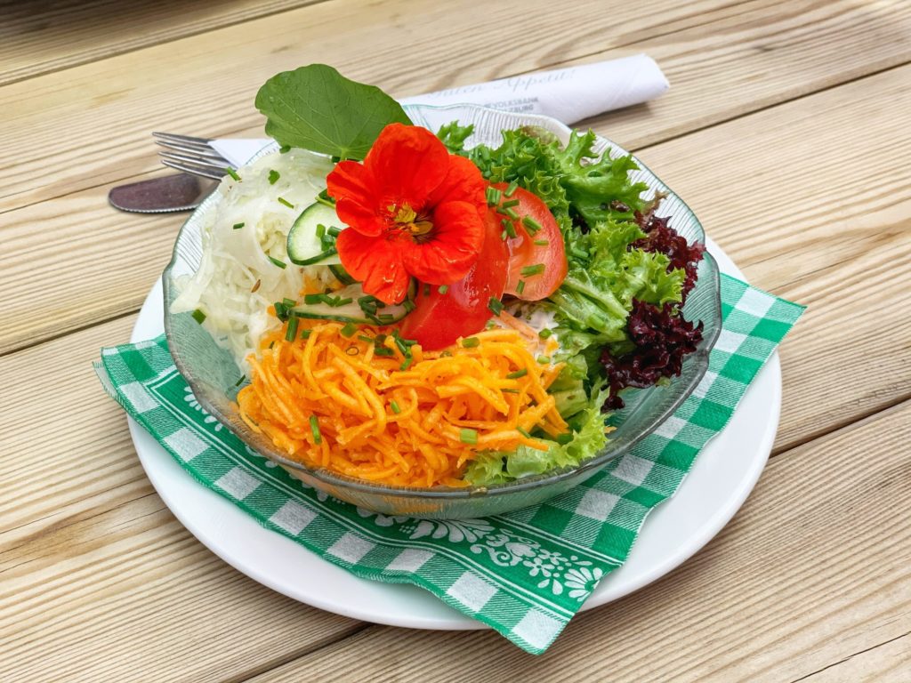 Das Foto zeigt eine Glasschüssel mit gemischtem Salat und einer roten Blüte darauf auf einer grün-karierten Serviette.