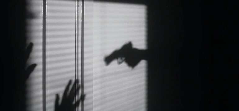 Eine Pistole ist auf eine Person gerichtet, die ihre Hände hochhält. Es sind jedoch nur die Schatten der beiden Menschen zu sehen, das Bild ist schwarz-weiß.