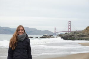 Die Stadt- und Umweltpsychologin Sandra Geiger steht am Strand, im Hintergrund ist das Meer sowie die Golden Gate Bridge zu sehen.