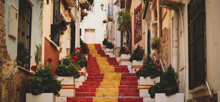 Eine Treppe in der Stadt Calp. Sie ist in den Farben der spanischen Flagge (rot, gelb, rot) angemalt. Links und rechts davon sind Häuser.