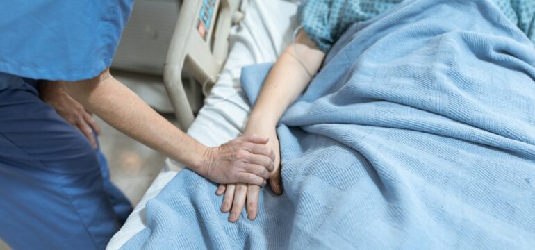 Eine Frau liegt in einem Krankenhausbett, ein Pfleger hält ihre Hand.