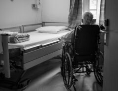 Ein älterer Mensch sitzt im Rollstuhl, vor ihm ist ein Bett zu sehen.