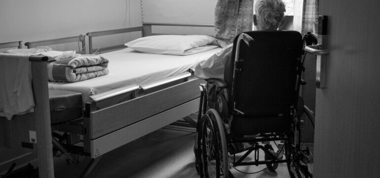 Ein älterer Mensch sitzt im Rollstuhl, vor ihm ist ein Bett zu sehen.