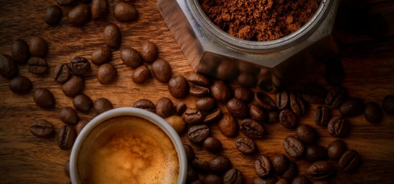 Eine Tasse Kaffee sowie ein Behälter mit gemahlenen Kaffee stehen auf einem Tisch, auf dem ebenso Kaffeebohnen liegen.