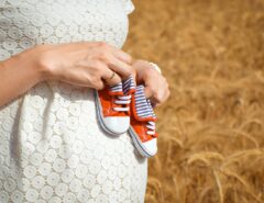 Eine Schwangere steht vor einem Feld und hält Babyschuhe gegen ihren Bauch.
