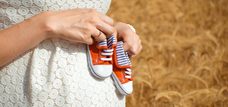 Eine Schwangere steht vor einem Feld und hält Babyschuhe gegen ihren Bauch.